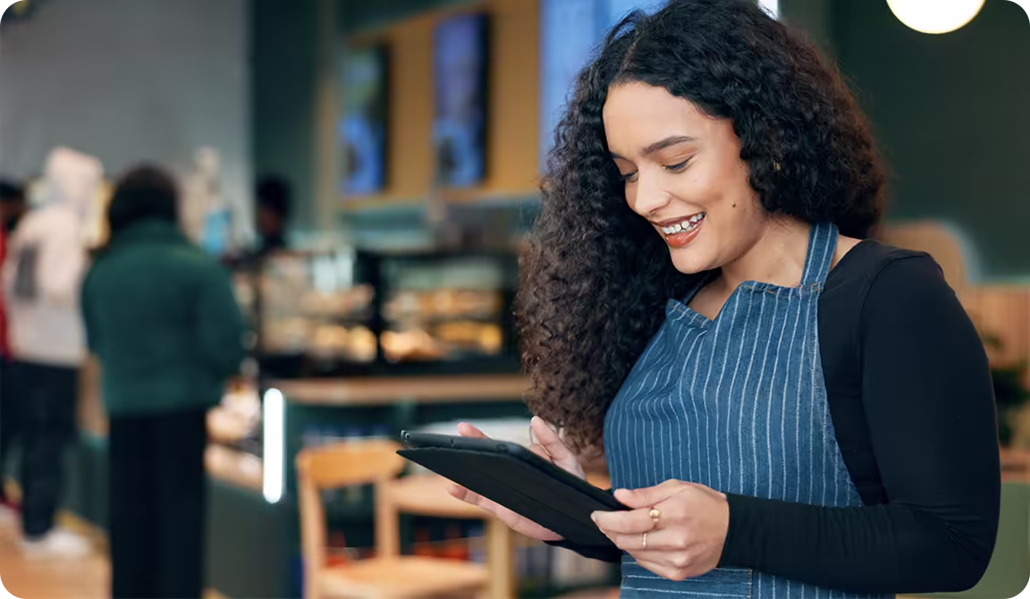 A smiling café worker in a striped apron uses a tablet.