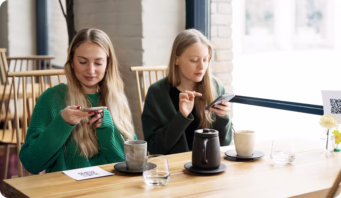 Two women use their smartphones at a café table with coffee, tea and flowers.